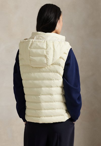 Femme de dos portant une doudoune crème à capuche par-dessus un haut à manches longues bleu marine et un pantalon foncé, sur un fond neutre.