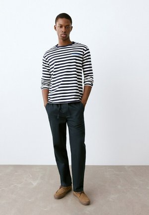 Giovane uomo in piedi con le mani nelle tasche, indossa una maglietta a maniche lunghe a righe bianche e nere, pantaloni neri e scarpe marroni, davanti a un muro bianco.