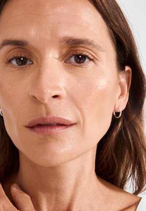 Nærbillede af en midaldrende kvinde med brune øjne, lys hud, iført små øreringe og en hvid top, der blidt rører ved sin nakke.