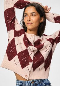 Gaiši rozā adīta jakas ar tumši sarkanu rombu rakstu, ribotu apakšu un trim pogām, ar tekstūrētu apdari un nedaudz īsu dizainu.