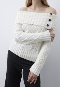 Cremefarbener, schulterfreier Pullover mit Zopfmuster, gerippten Bündchen und einem breiten Ausschnitt mit drei dunklen Knöpfen auf der linken Seite.