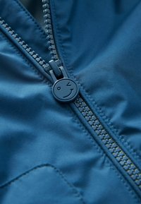 Blauwe puffy jassectie met een gladde stof, zichtbare stiksels en een rits met een smiley-gezicht ontwerp.