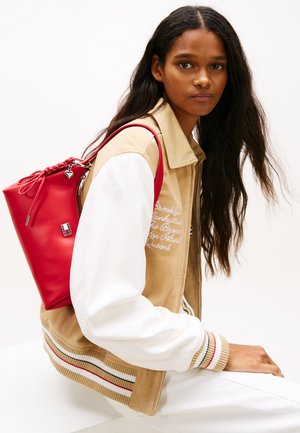 Ženska z dolgimi temnimi lasmi, oblečena v bež in belo jakno ter bele hlače, sedi in nosi rdečo torbo z vrvico čez ramo.