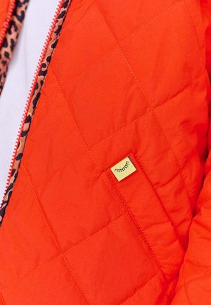 Převlečená oranžová bunda s prošívaným vzorem, která má podšívku s leopardím potiskem a malou žlutou visačku s jednoduchým designem obličeje poblíž kapsy.
