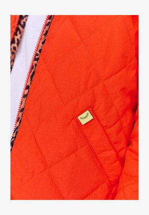 Převlečená oranžová bunda s prošívaným vzorem, která má podšívku s leopardím potiskem a malou žlutou visačku s jednoduchým designem obličeje poblíž kapsy.