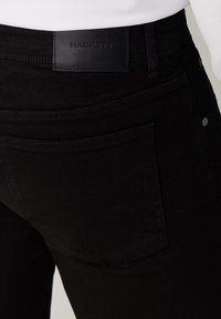 Vaqueros negros de denim con un diseño ajustado, que presentan una etiqueta de cuero con la marca "HACKETT", costuras visibles y una disposición clásica de cinco bolsillos.