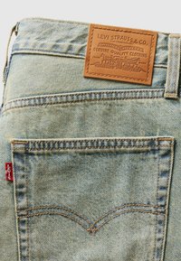 Svijetloplave traperice s tamnoplavim kožnim etiketom označenom "Levi Strauss & Co." i crvenom etiketom sa strane, s istaknutim šavovima.