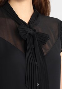 Blusa nera con una parte superiore trasparente e un dettaglio a fiocco annodato sul colletto. Il tessuto appare liscio con accenti plissettati.
