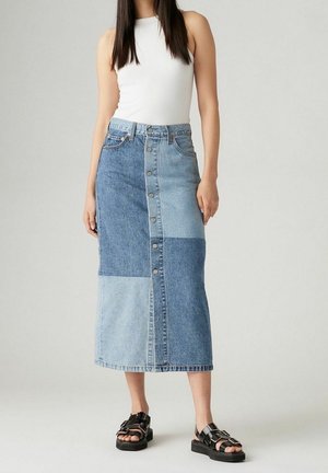 Kvinde iført hvid ærmeløs top, patchwork denim midinederdel med knapper foran og sorte sandaler med spænde, der står mod en ensfarvet baggrund.