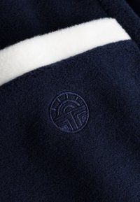 Navy fleece fabric with a white interior lining; features a circular embroidered logo in dark navy on the front. Textured, soft appearance.