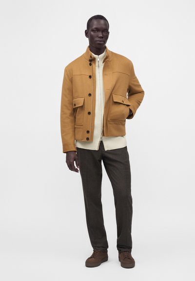 Homme portant une veste couleur moutarde par-dessus un pull blanc zippé, un pantalon sombre et des chaussures marron, debout avec une main dans la poche de la veste.