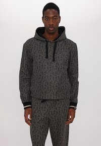 Mikina s kapucí a odpovídající kalhoty v černé barvě s šedým opakujícím se textovým vzorem. Obsahuje žebrované manžety a pás.