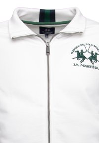 White zip-up jacket with a high collar, featuring green logo embroidery and a striped green and black collar. Smooth cotton material.