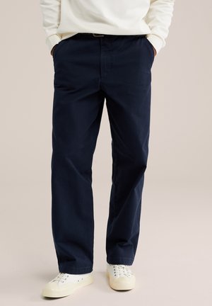 Homme portant un pantalon droit bleu marine, des baskets blanches et un haut crème à manches longues, les mains dans les poches.