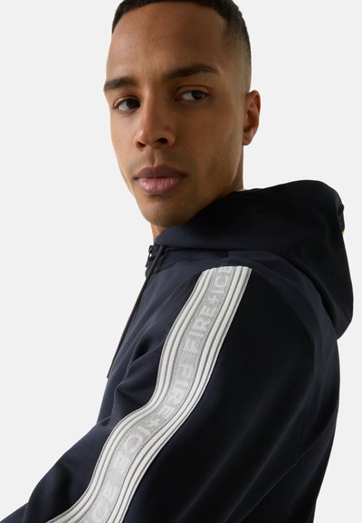 Marineblaues Hoodie mit Frontreißverschluss, Kängurutasche und kontrastierenden weißen Streifen mit "FIRE"-Muster an den Ärmeln. Glatte Stoffoberfläche.