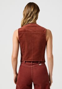 Vesta din corduroy de culoare ruginită, cu un design scurt, fără mâneci și detalii vizibile ale cusăturii. Țesătură texturată și branding subtil pe spate.