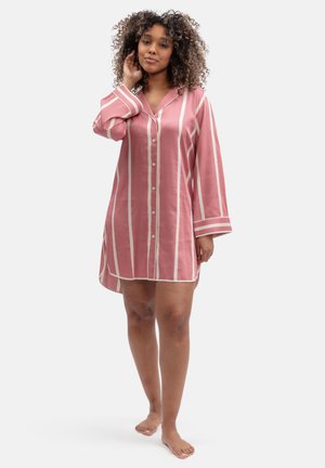 Mujer con cabello rizado, vistiendo una camisa de dormir de manga larga con botones y rayas verticales rosas y blancas, de pie descalza sobre un fondo liso.