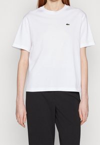 Bílá bavlněná tričko s krátkými rukávy, klasickým dekoltem a zeleným vyšitým logem na levé hrudi. Elegantní, minimalistický design.