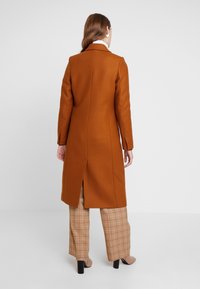 Long brown coat with a tailored design, split hem, and shoulder pads, paired with beige checkered trousers and beige shoes.