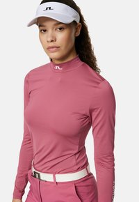 Långärmad rosa pikétröja med hög krage och logotyp vid nacken, tillsammans med vit bälte. Slät, elastisk tyg, figursydd passform.