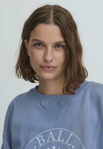 Zila sporta krekla ar ribotu apkakli un lielu apaļu grafiku ar vārdu "BALL" baltā krāsā, kurai pievienoti zvaigznes un gluda tekstūra.