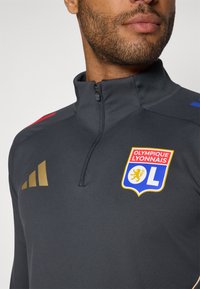 Grå atletisk jakke med lynlås krave, med guld Adidas-logo, røde og blå accenter, samt Olympique Lyonnais-emblemet på brystet.