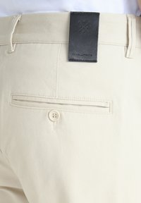 Beżowe bawełniane spodnie z tylną kieszenią zapinaną na guzik oraz czarną skórzaną metką z wytłoczonym logo nad pasem.