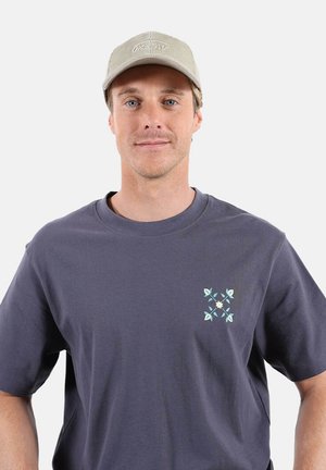 Jeune homme portant une casquette beige et un t-shirt gris foncé avec un petit motif floral bleu et blanc sur la poitrine, debout devant un fond uni.
