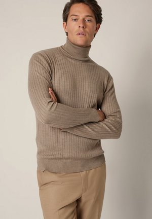 Mężczyzna o jasnobrązowych włosach, ubrany w beżowy prążkowany golf i beżowe spodnie, stojący z założonymi rękami na tle gładkiej ściany.