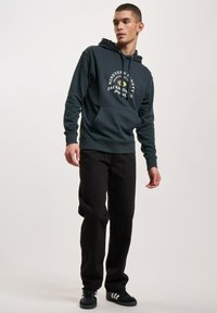 Dunkelgrüne Kapuzenjacke mit weißem Grafikdruck und Kordelzugkapuze, kombiniert mit schwarzen Hosen und schwarzen Sneakers mit weißen Streifen.