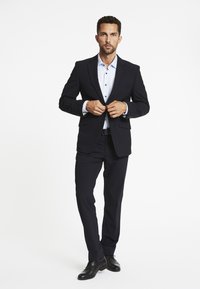 Man dichtknopen van de zwarte colbert, gekleed in een lichtblauw overhemd, zwarte broek en zwarte nette schoenen, staand op een witte achtergrond.