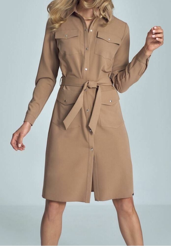 Femme portant une robe-chemise beige mi-longue à manches longues, avec des poches avant et une ceinture nouée à la taille, debout devant un fond uni.