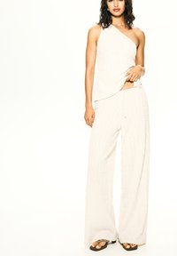 Haut tricoté blanc à une épaule, associé à un pantalon large à texture et coupe ample, avec des tongs marron et une taille élastique à cordon.
