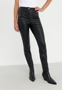 Pantalons en simili cuir noir avec un design ajusté, présentant deux poches zippées à l'avant, une taille haute et des jambes fuselées. Assortis avec des bottes à talons noires.