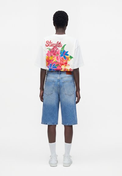 Άτομο με την πλάτη στραμμένη φοράει λευκό T-shirt με πολύχρωμο floral σχέδιο και το κείμενο "Steglitz", τζιν σορτς μήκους έως το γόνατο, λευκές κάλτσες και αθλητικά παπούτσια.
