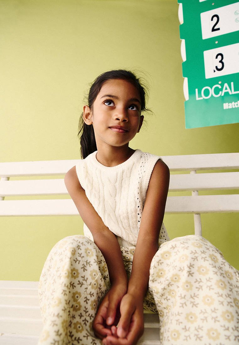Jeune fille en haut blanc en maille sans manches et pantalon fleuri, assise sur un banc blanc, regardant vers le haut avec un doux sourire contre un mur vert clair.