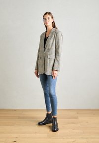 Femme debout sur un sol en bois, portant un blazer à carreaux, un haut noir, un jean skinny bleu, des bottines noires et des lunettes transparentes, devant un mur gris uni.