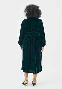 Grünes Samtkleid mit langen Ärmeln und tailliertem Bund, das eine glatte Textur und einen bodenlangen Schnitt aufweist, von hinten betrachtet.