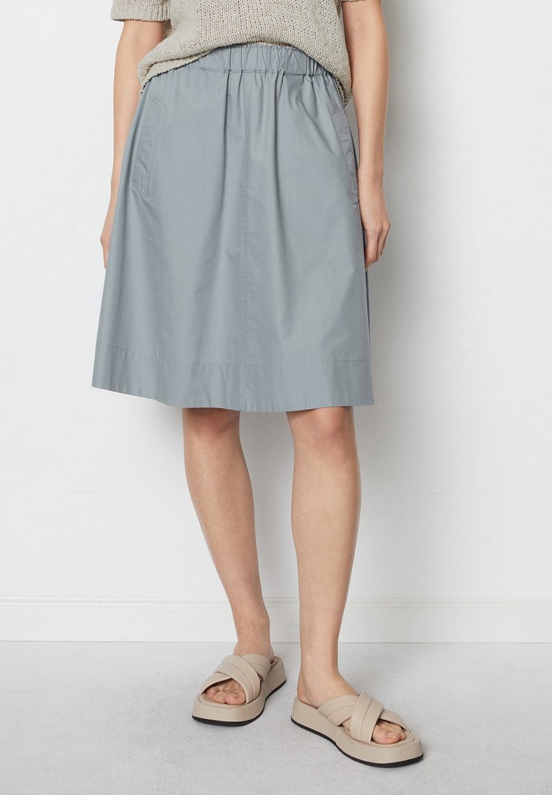 Femme portant une jupe gris clair mi-genoux, un haut beige en maille et des sandales beige à lanières croisées, debout contre un mur blanc.