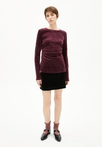 Plüschiges, tief burgunderrotes langärmliges Oberteil mit gerafftem Bund, kombiniert mit einem schwarzen Minirock. Das Model trägt gemusterte Socken und schwarze Schuhe.