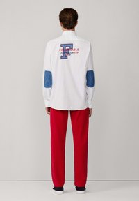 Chemise blanche à manches longues avec des coudières en denim bleu, des graphiques brodés et des lettres rouges, assortie à un pantalon rouge et des chaussures foncées.