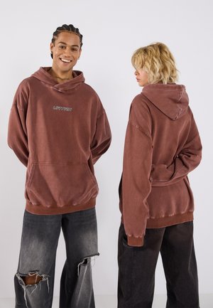Overdimensjonert, rosa-brun hettegenser med frontlomme, snor i hetta og brodert tekst. Kombinert med slitte svarte vide jeans.
