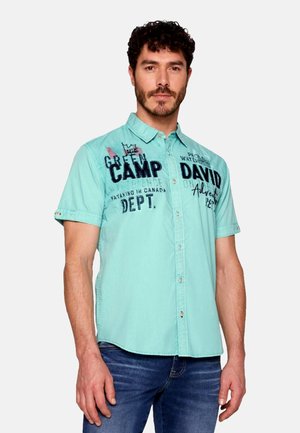 Camicia azzurra a maniche corte con chiusura a bottoni e grafiche testuali in blu scuro. Realizzata in tessuto dalla texture liscia, presenta un colletto a punta.