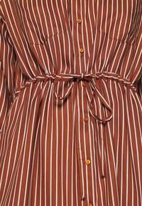 Chemise rayée brune avec des lignes verticales blanches, dotée d'un devant à boutonnière, d'une taille à cordon de serrage et de boutons orange. Texture douce.