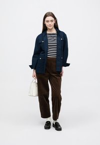 Chaqueta utilitaria azul marino sobre una camisa de rayas blancas y negras, pantalones de pana marrones, bolso blanco y mocasines negros de diseño elegante.