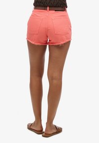 Pantalones cortos de mezclilla coral con dobladillo desflecado y dos bolsillos traseros, combinados con sandalias marrones, vistos desde atrás. Costuras detalladas visibles.