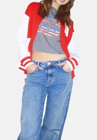 Une veste varsity rouge et blanche sur un t-shirt court gris avec un imprimé graphique. Un jean bleu clair taille haute avec une coupe décontractée.