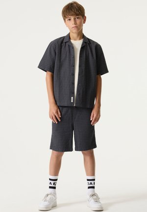 Chemise à manches courtes gris foncé avec un motif texturé, fermeture à boutons, associée à un short assorti. Chaussettes blanches montantes et chaussures de sport complètent le look.