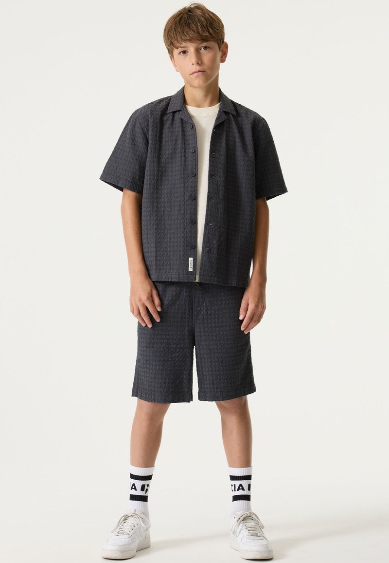 Chemise à manches courtes gris foncé avec un motif texturé, fermeture à boutons, associée à un short assorti. Chaussettes blanches montantes et chaussures de sport complètent le look.