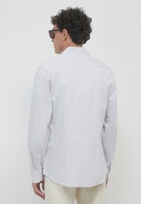 Uomo con capelli ricci che indossa occhiali da sole neri, una camicia a maniche lunghe grigia chiara a righe e pantaloni beige, di spalle su sfondo bianco.